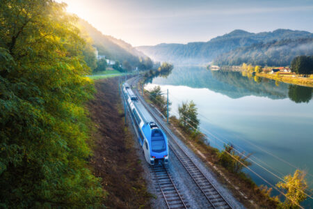 Aerial View Of Blue Modern High Speed Train Moving Near River In Alpine Mountains In Fog At Sunrise In Autumn. Top View Of Train, Railroad, Lake, Reflection, Trees In Fall. Railway Station In Slovenia